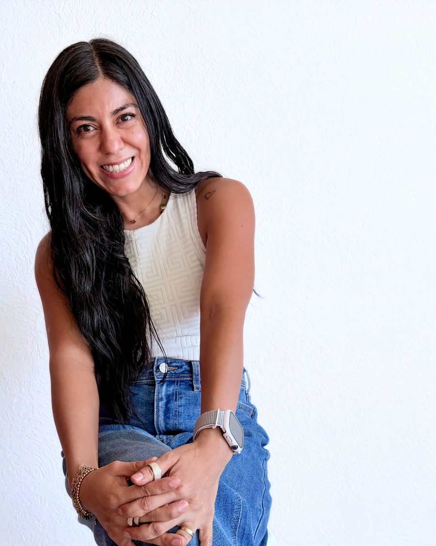Mujer sonriendo, sentada con jeans y top blanco frente a una pared clara